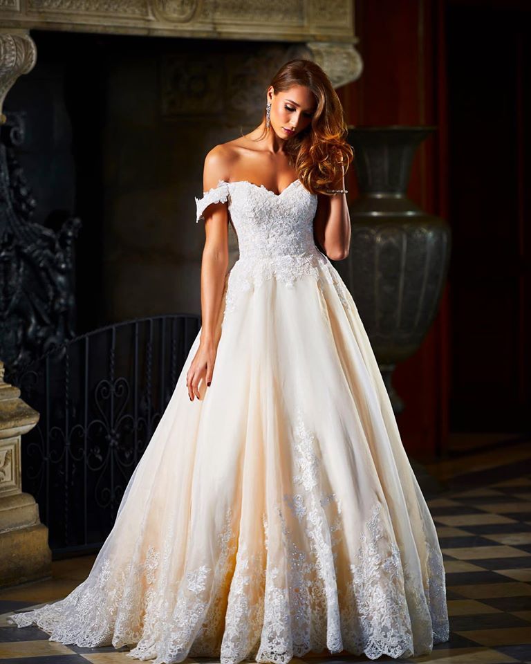Woman in a white off-the-shoulder wedding gown, standing in a room with a decorative fireplace and large vase.