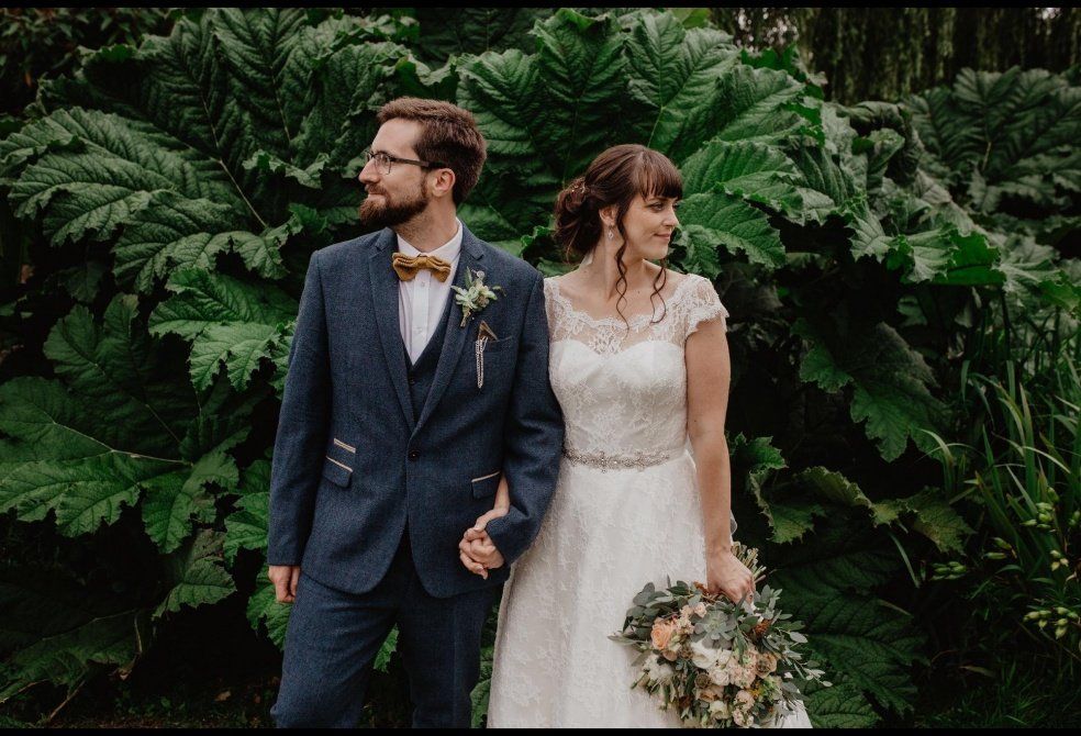 Couple in wedding attire posing by large green foliage. Groom in blue suit, bride in white dress, holding bouquet.