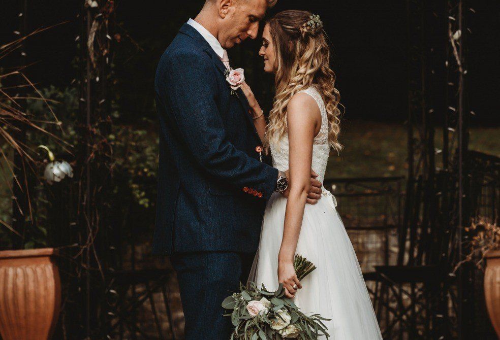 Bride and groom embracing, wedding day. She wears a white dress; he a blue suit. They are outdoors near potted plants.