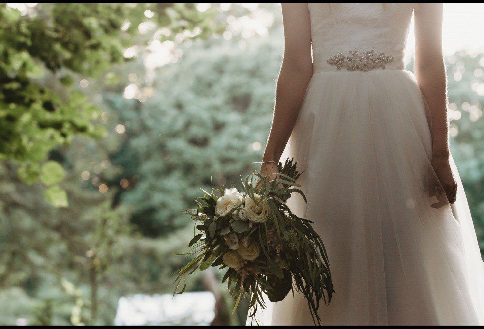 Bride in white dress holds bouquet, standing outdoors with sunlight in the background.
