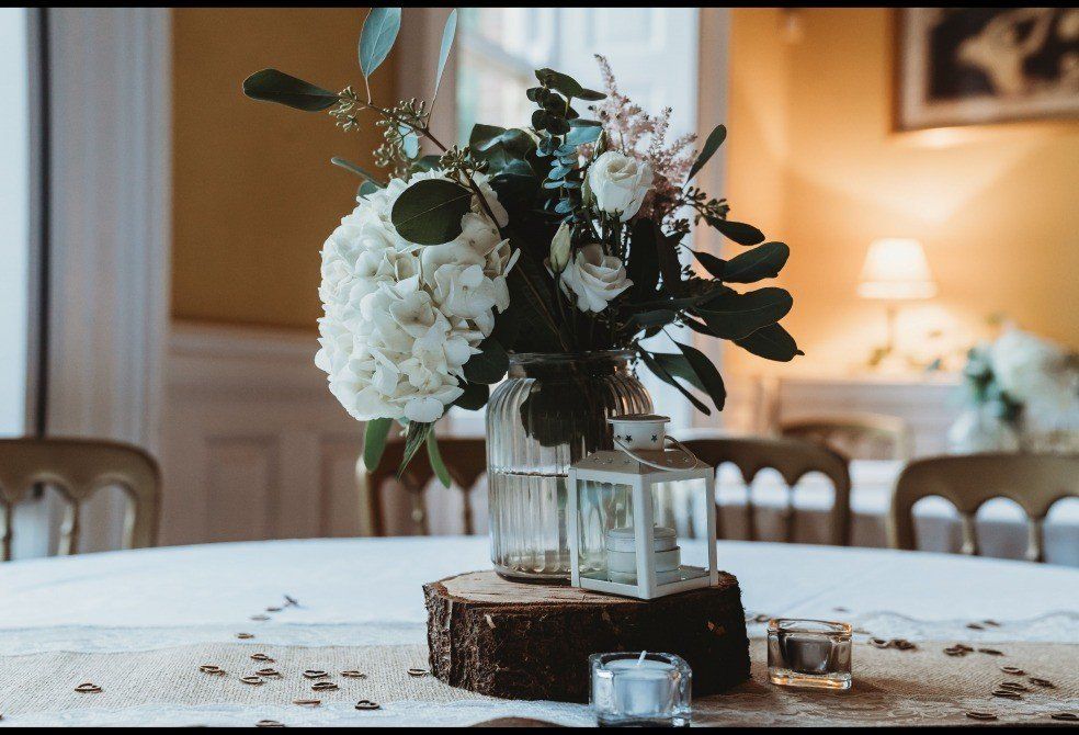 Centerpiece with white flowers, lantern, and candles on a wooden base on a decorated table.