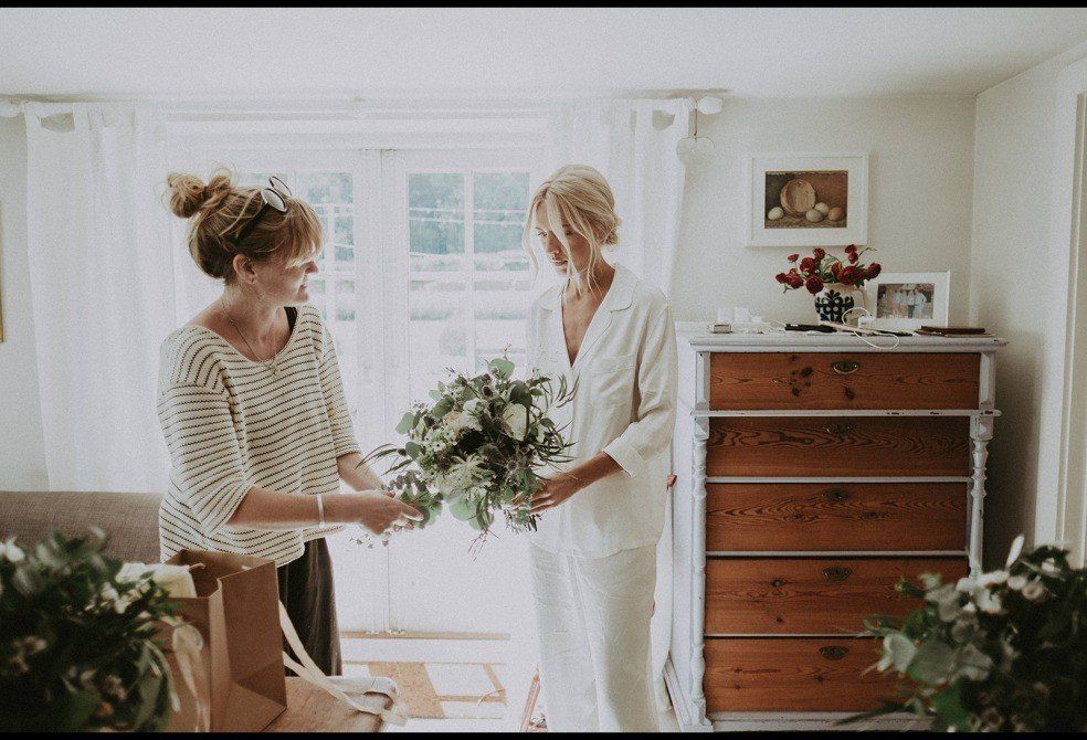 Woman in white pajamas receiving a bouquet from another woman in a striped shirt, indoors with bright light.