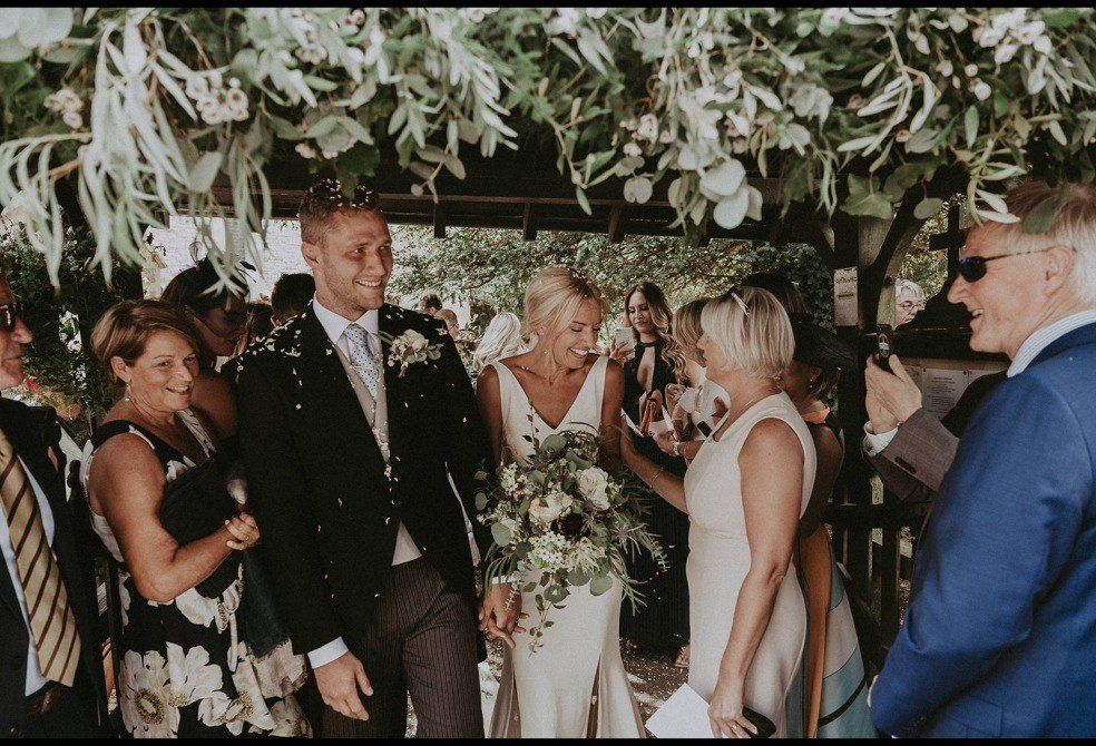Newlyweds showered with confetti under greenery. Guests smile.