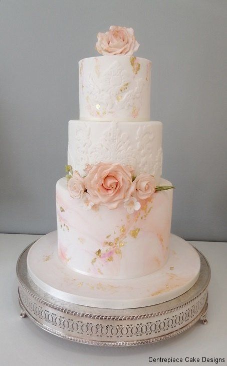 Three-tiered wedding cake, off-white with peach and gold accents, roses, on a silver cake stand.