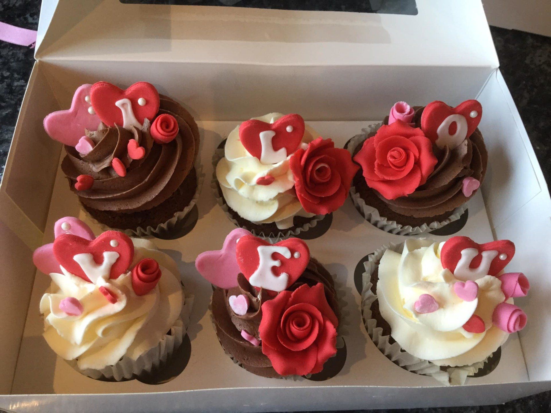 Six Valentine's cupcakes in a box, decorated with red roses, heart toppers, and 
