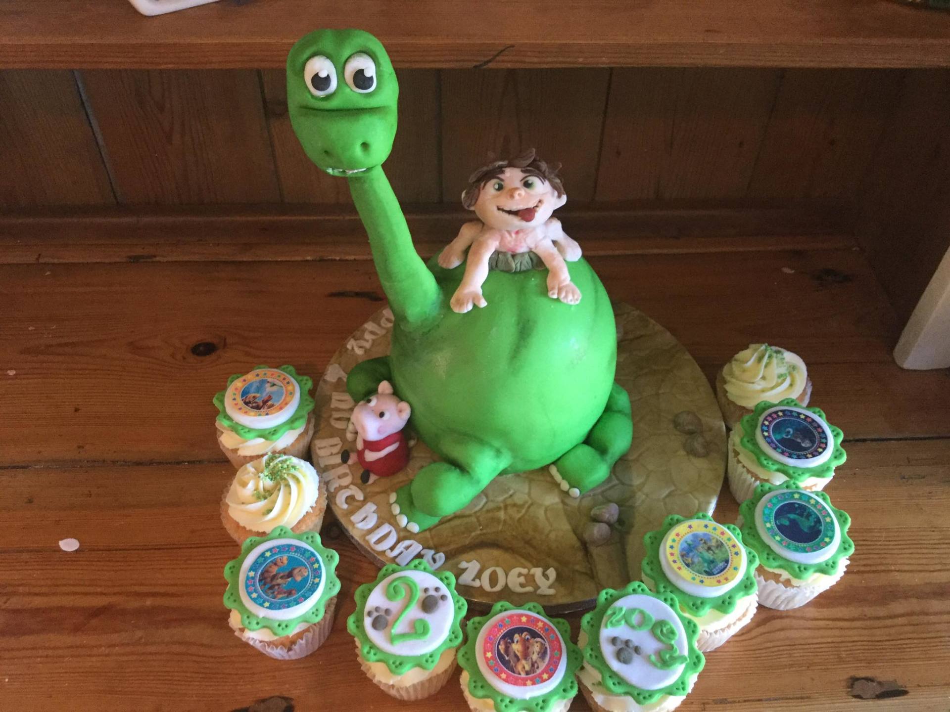 Cake decorated as a green dinosaur with a boy on its back, surrounded by cupcakes.