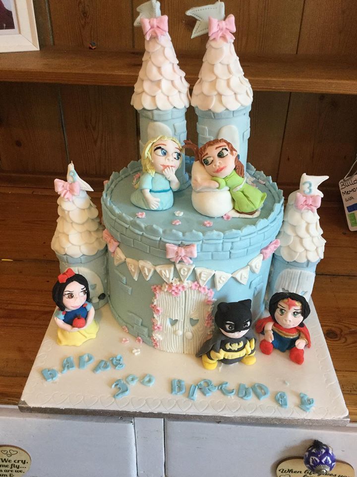 A decorated castle cake featuring cartoon princess and superhero figurines, light blue frosting, and floral accents.