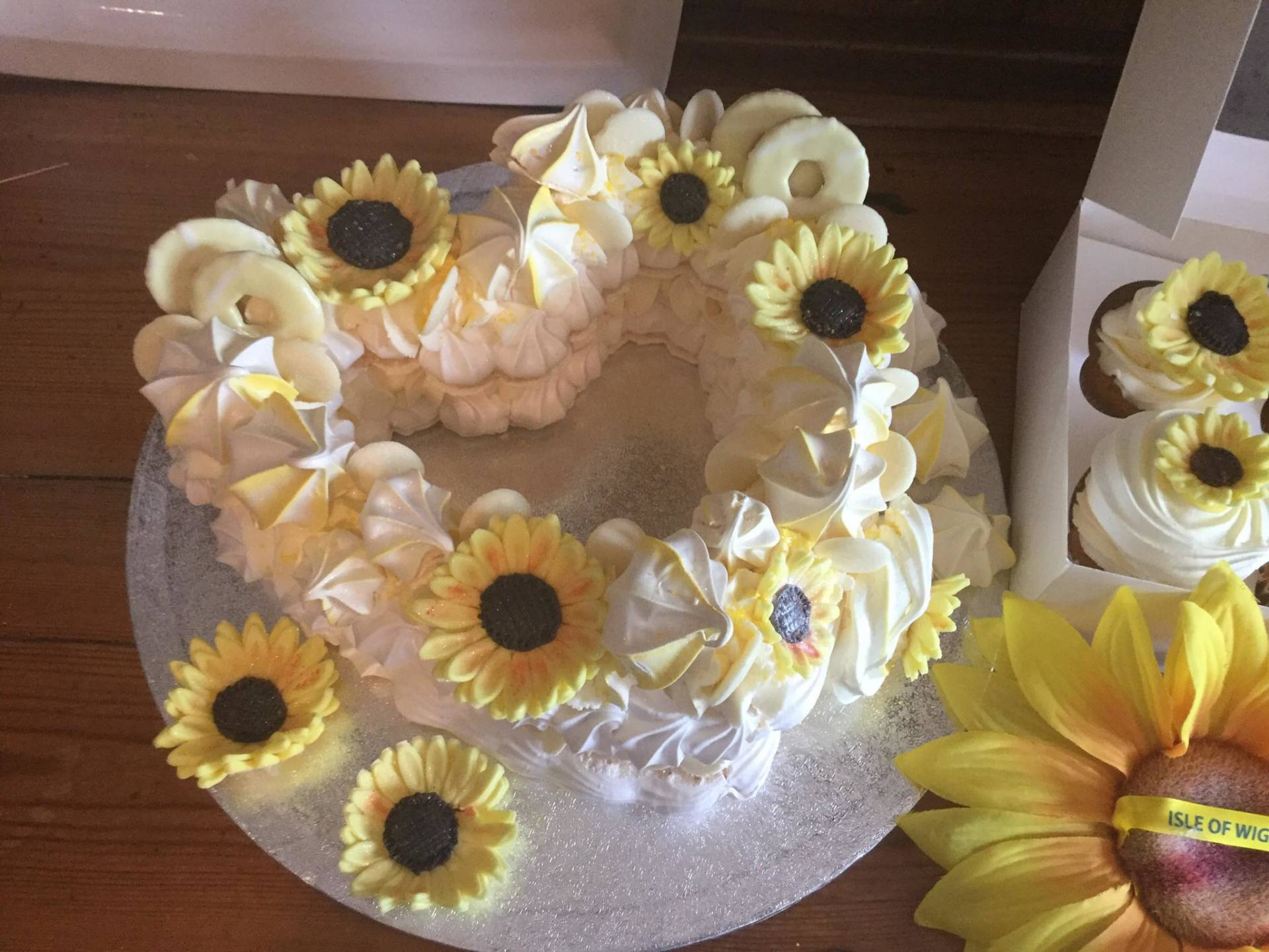 Heart-shaped cake decorated with white frosting, yellow sunflowers, and white chocolate rings. Cupcakes and a sunflower are nearby.