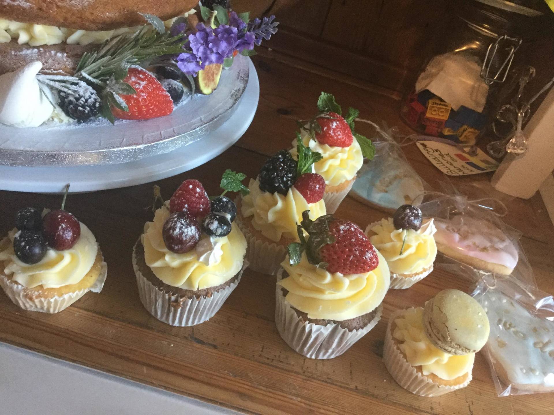 Cupcakes with yellow frosting and berries on a wooden surface, with a pastry plate and jar in background.