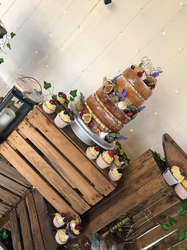 Naked wedding cake with fruit and cupcakes on wooden crates, decorated with string lights and greenery.