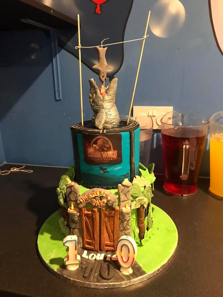 Dinosaur-themed birthday cake with a gate, logo, and figurine, teal and green.
