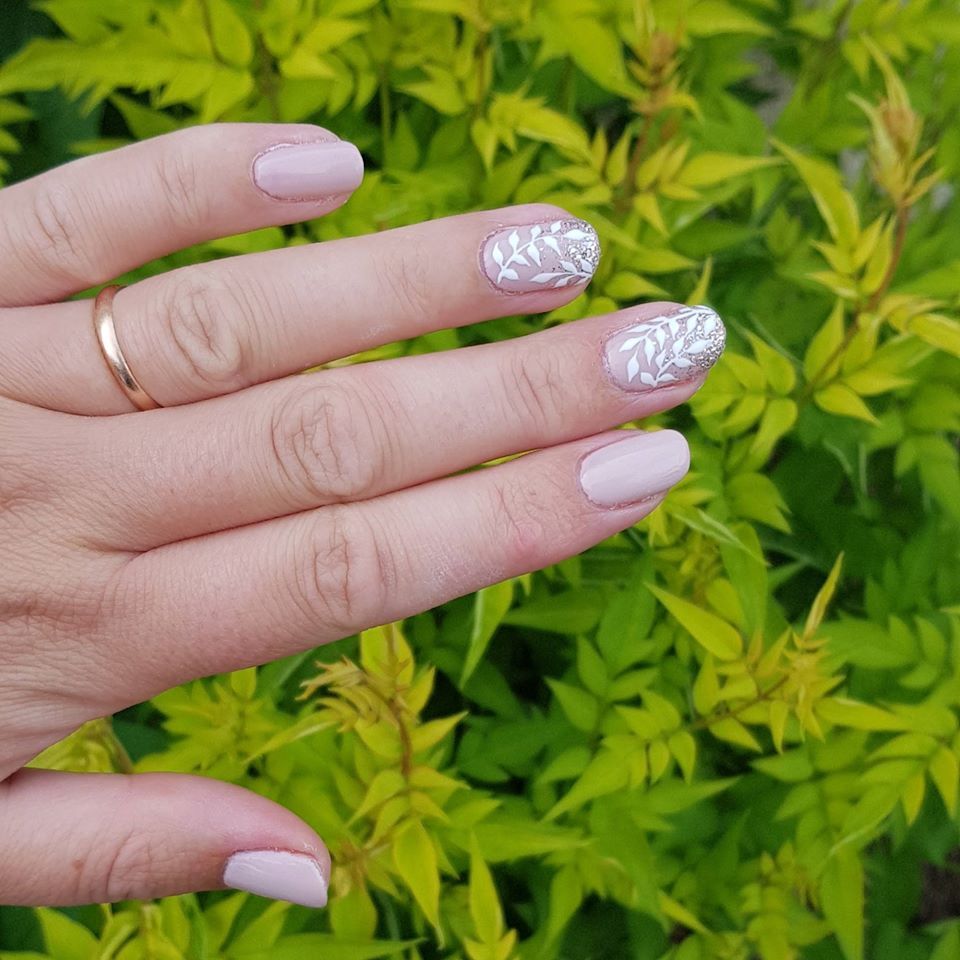 Hand with light pink nails, two decorated with white leaf designs, resting against green foliage.