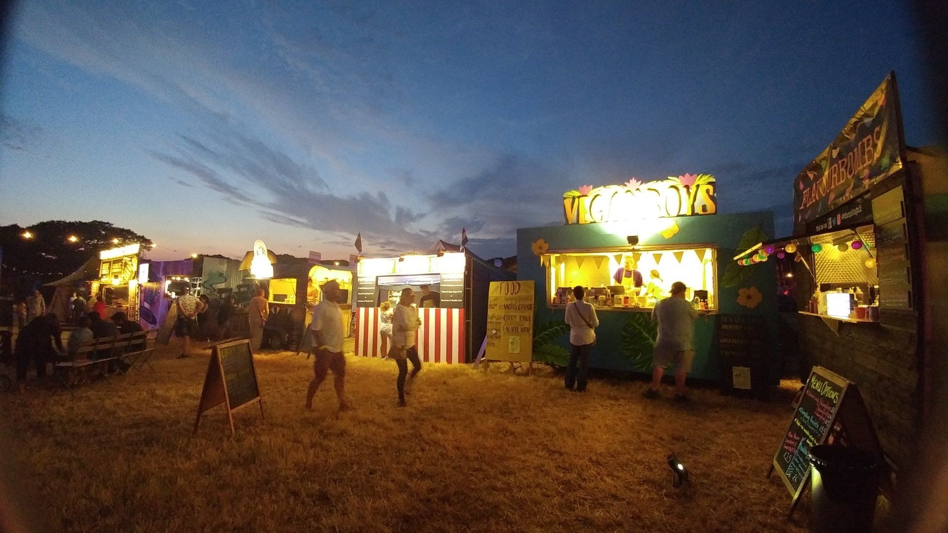 Food stalls at dusk, illuminated by lights; people gather and walk around.