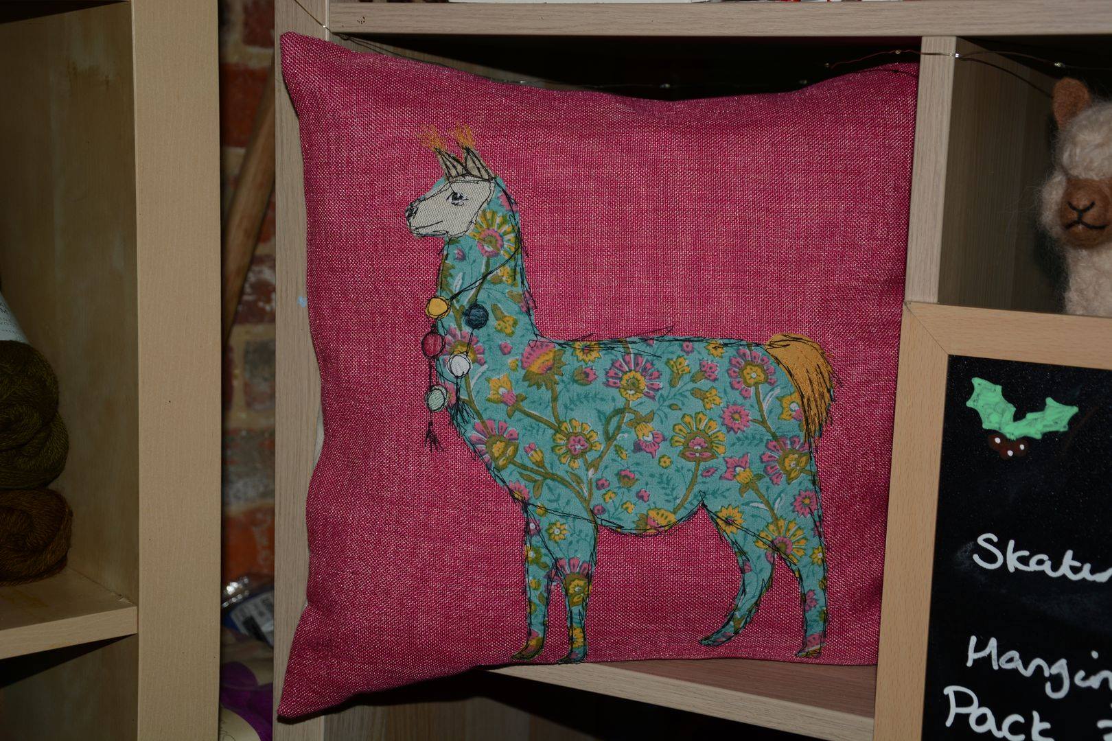 Pink pillow with llama design in a shop setting. Llama is decorated with colorful floral patterns and lights.