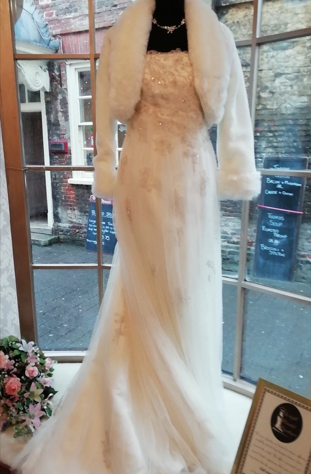 Wedding dress on display, ivory with gold detailing, paired with a white fur bolero, showcased in a window.