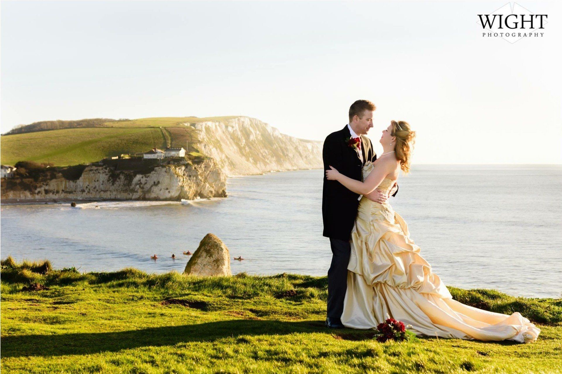 Couple embracing on a grassy cliff overlooking a blue ocean; wedding dress, suit, and scenic coastal view.