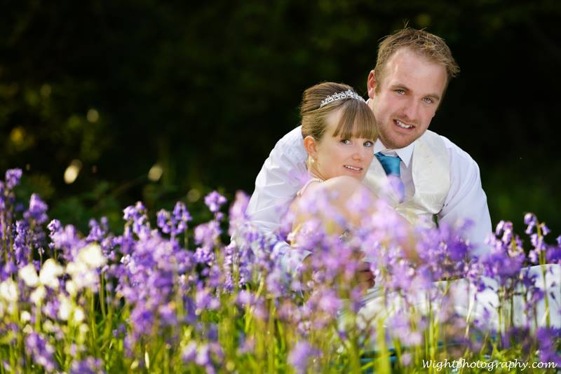 Couple in wedding attire embrace in a field of purple flowers; man behind woman with a slight smile.