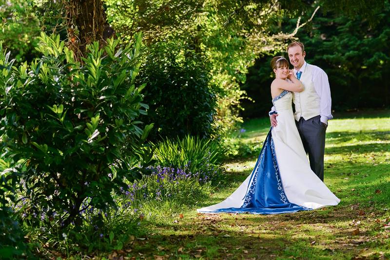 Couple in wedding attire, embracing on a grassy lawn with trees in the background. She wears a blue and white gown.