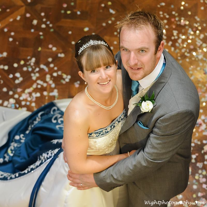 Newlyweds embrace, confetti on the floor. Bride wears white and blue gown, tiara. Groom in gray suit, turquoise tie.