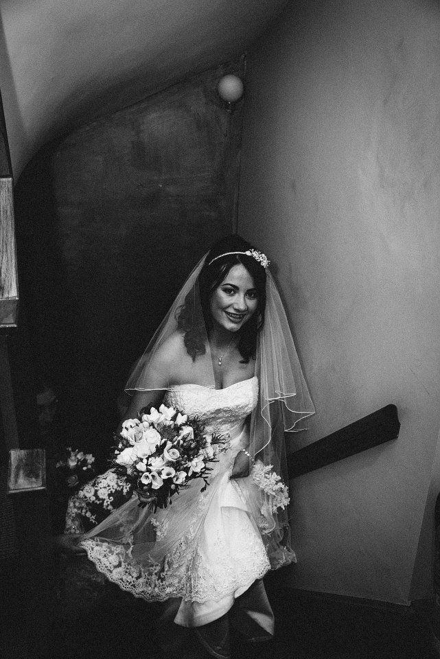 Bride in a white dress, veil, and holding a bouquet, descending a dark staircase.