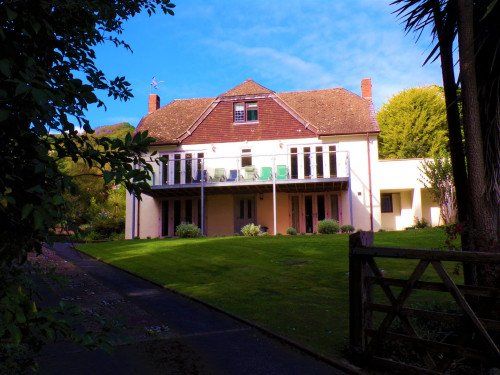 Two-story beige house with red roof and balcony, set on a green lawn with a driveway and trees.