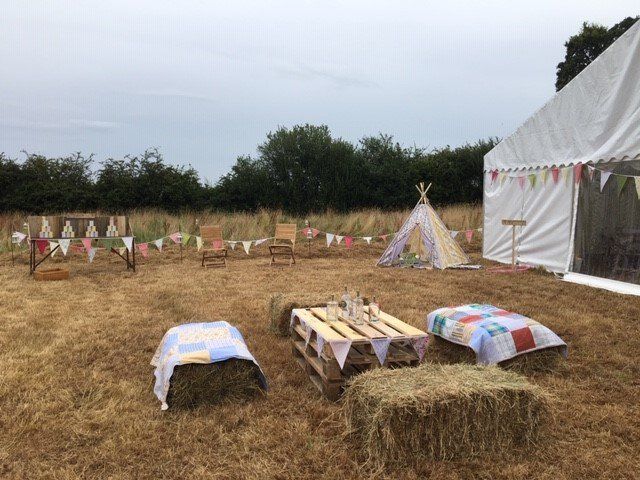 Outdoor party setup: tent, teepee, hay bale seating, wooden pallet table, and bunting in a field.