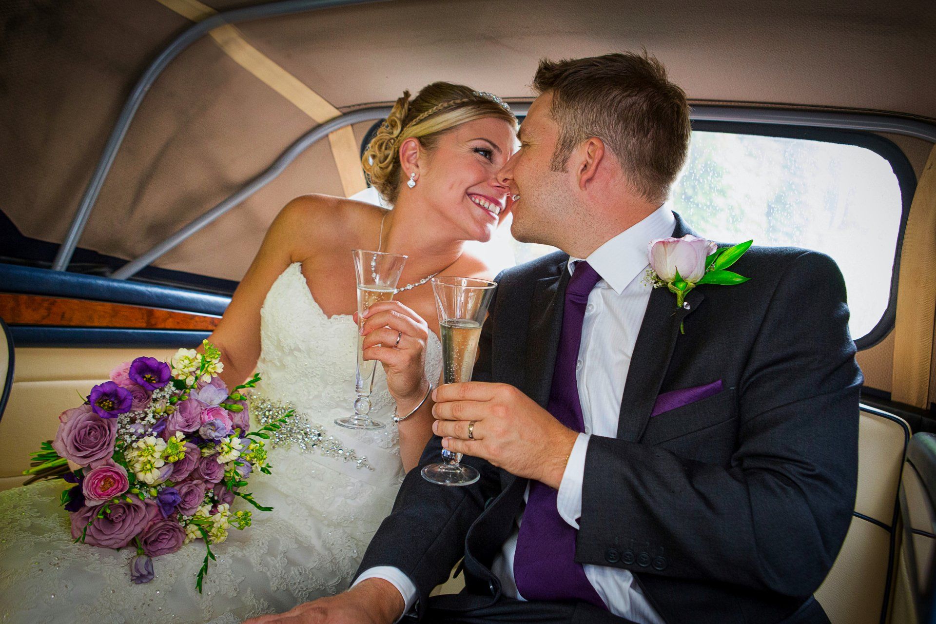 Bride and groom toast with champagne in a vintage car; she wears a white gown, he wears a suit.