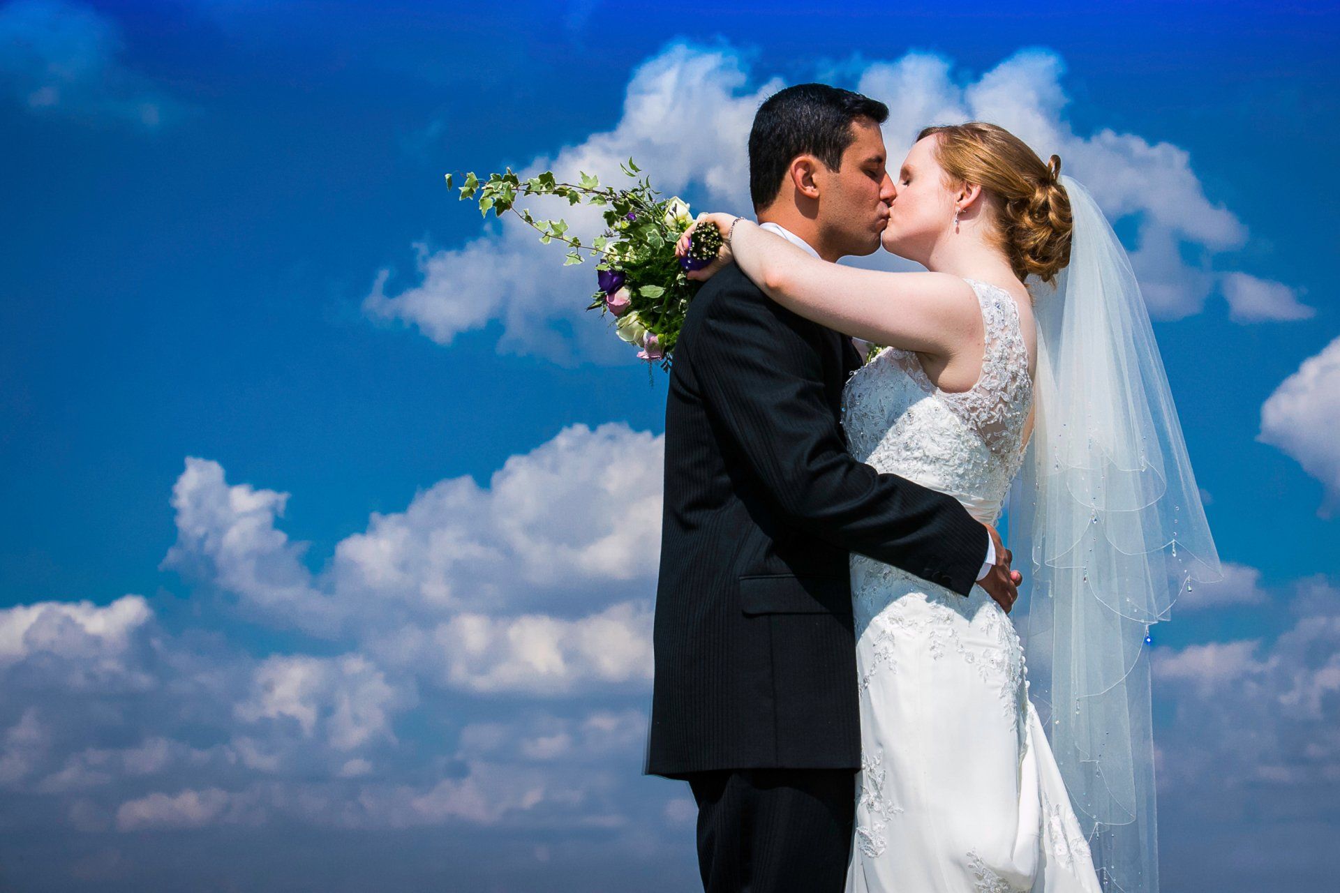 Couple in wedding attire kissing, blue sky and clouds in background.