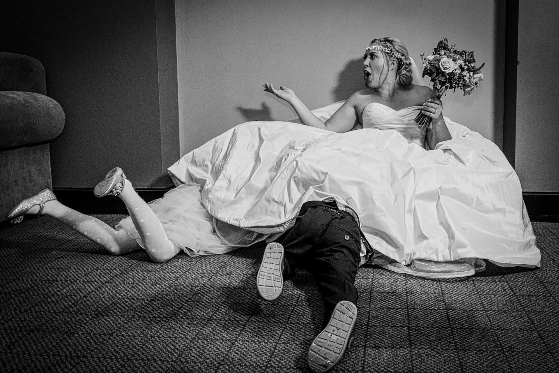 Bride and child playfully wrestling under her wedding dress; bride laughs, holding bouquet.