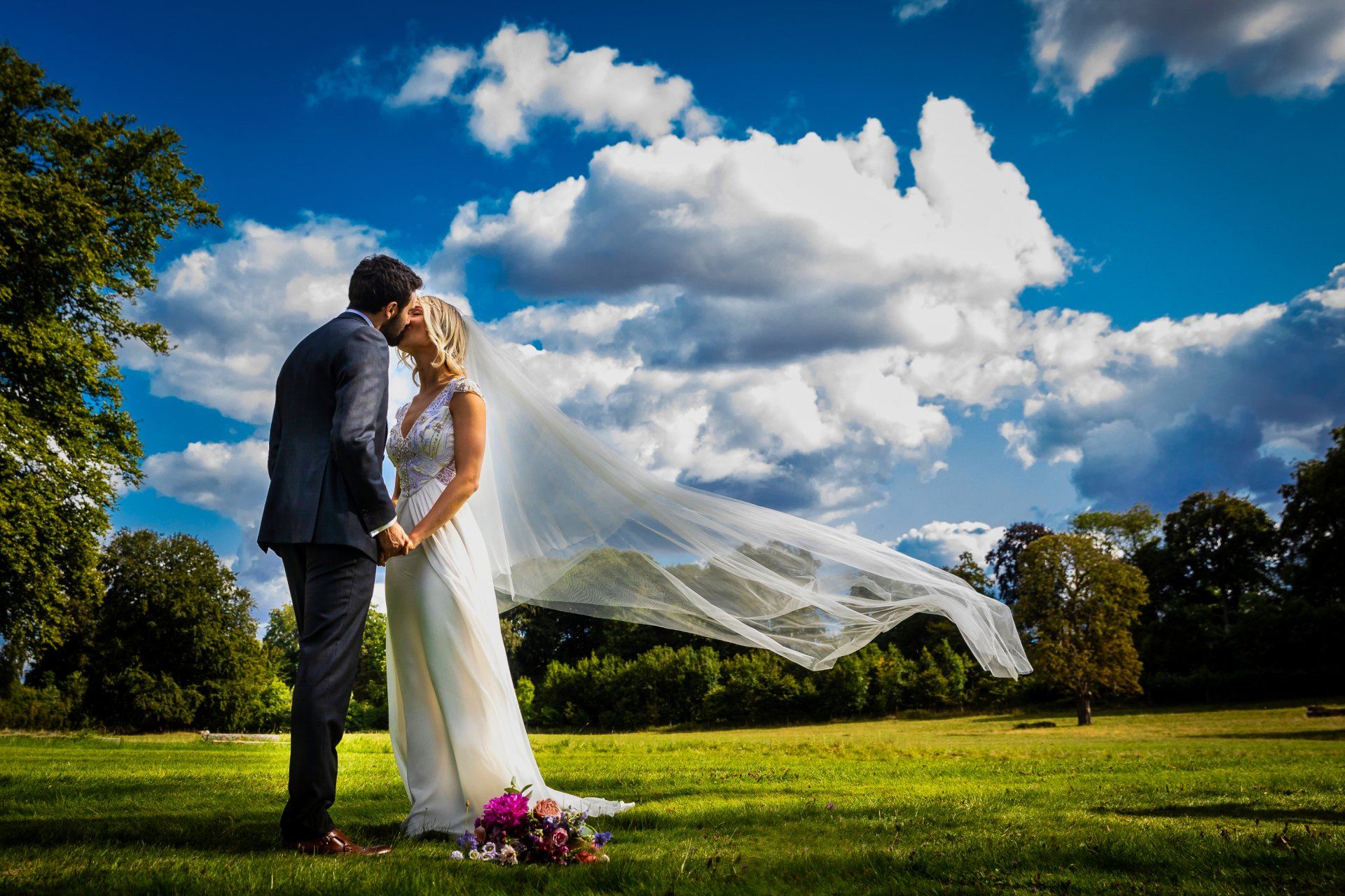 Couple kissing, bride's veil billowing in the wind, holding hands on grassy field, blue sky with puffy white clouds.