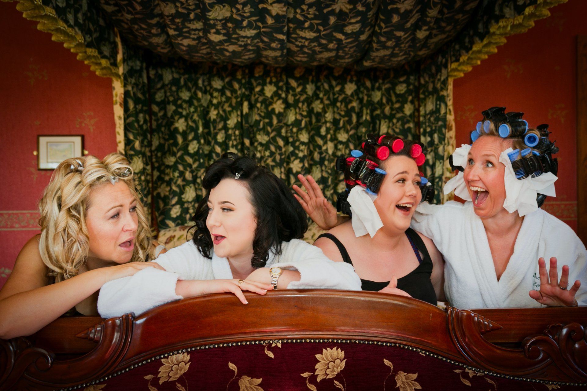 Four women in robes and curlers laugh on a bed with red walls and a decorated headboard.