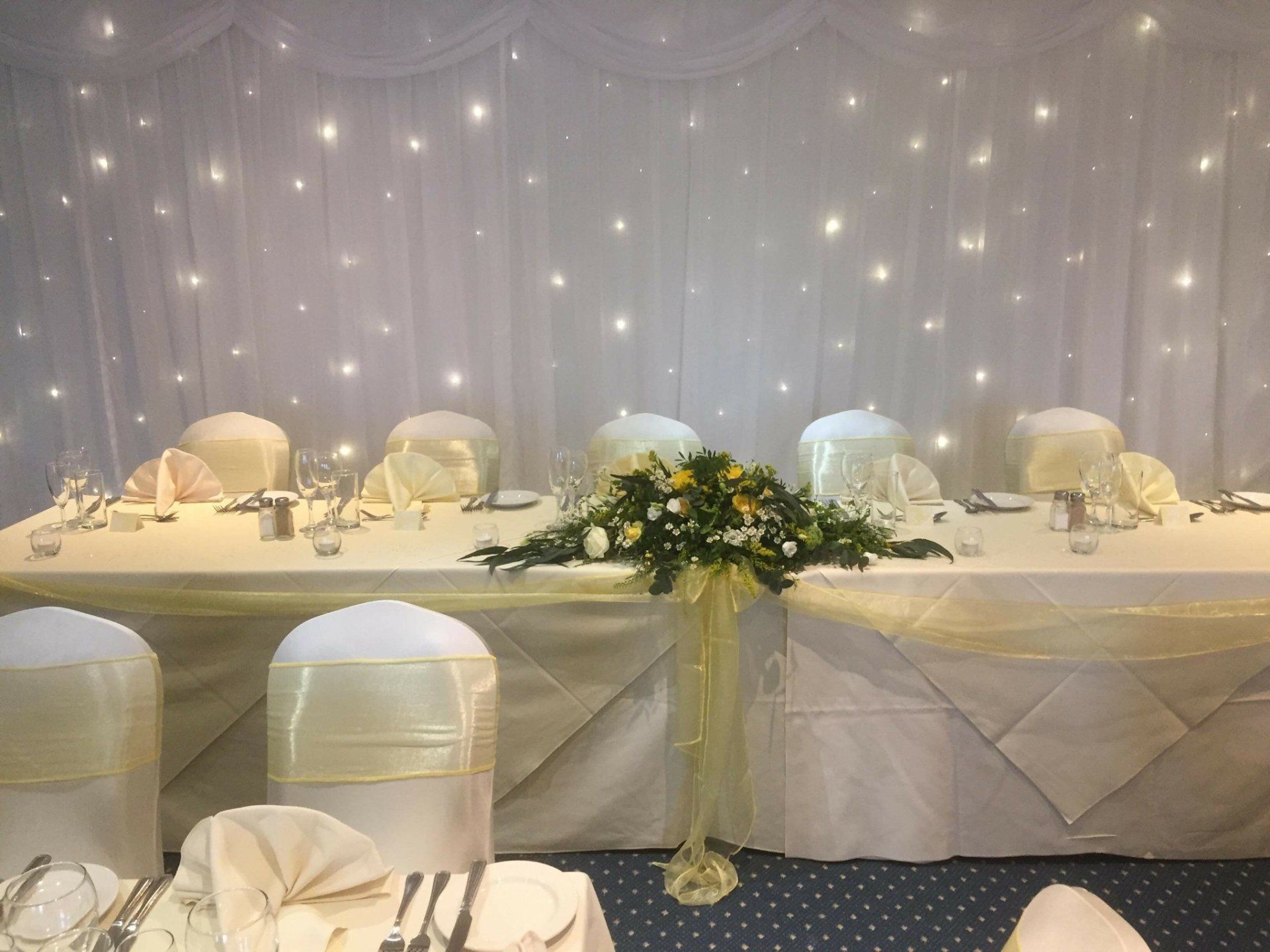 Wedding reception table with floral centerpiece, cream linens, and twinkling light backdrop.