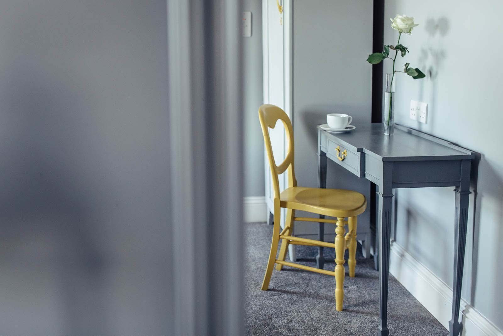 Yellow chair at a desk with a white rose in a vase; room with light gray walls.