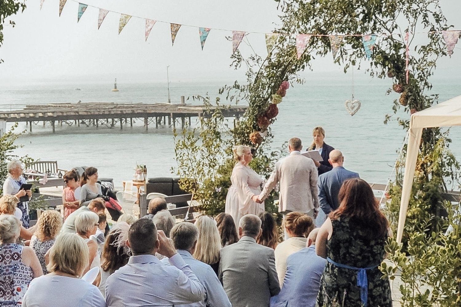 Wedding ceremony by the sea, with couple holding hands under floral arch, guests seated.