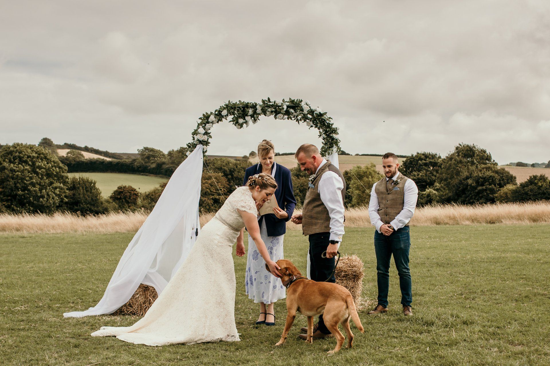 Bride petting a dog at outdoor wedding ceremony; officiant and groom watch. Green fields, floral arch.