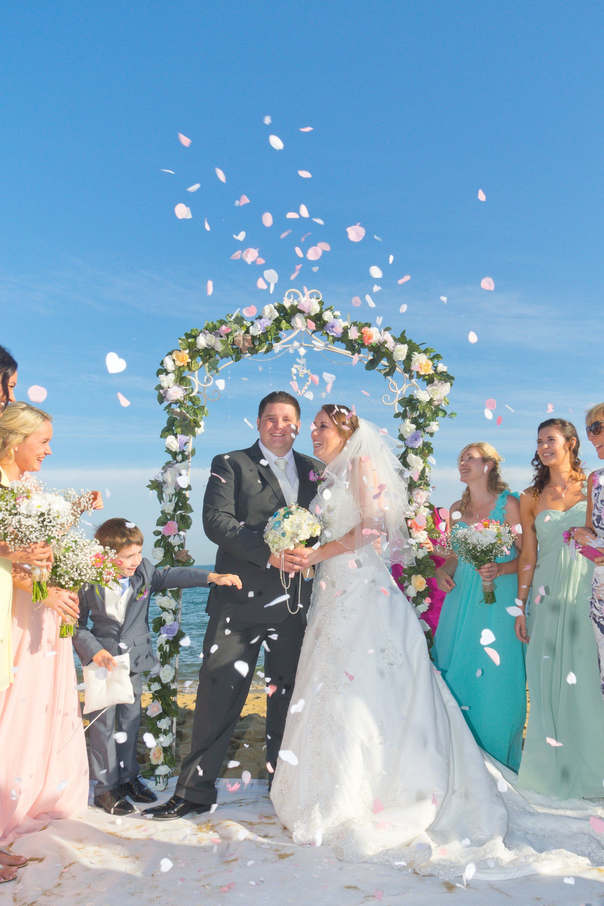 Bride and groom under floral arch, showered with petals at beach wedding.