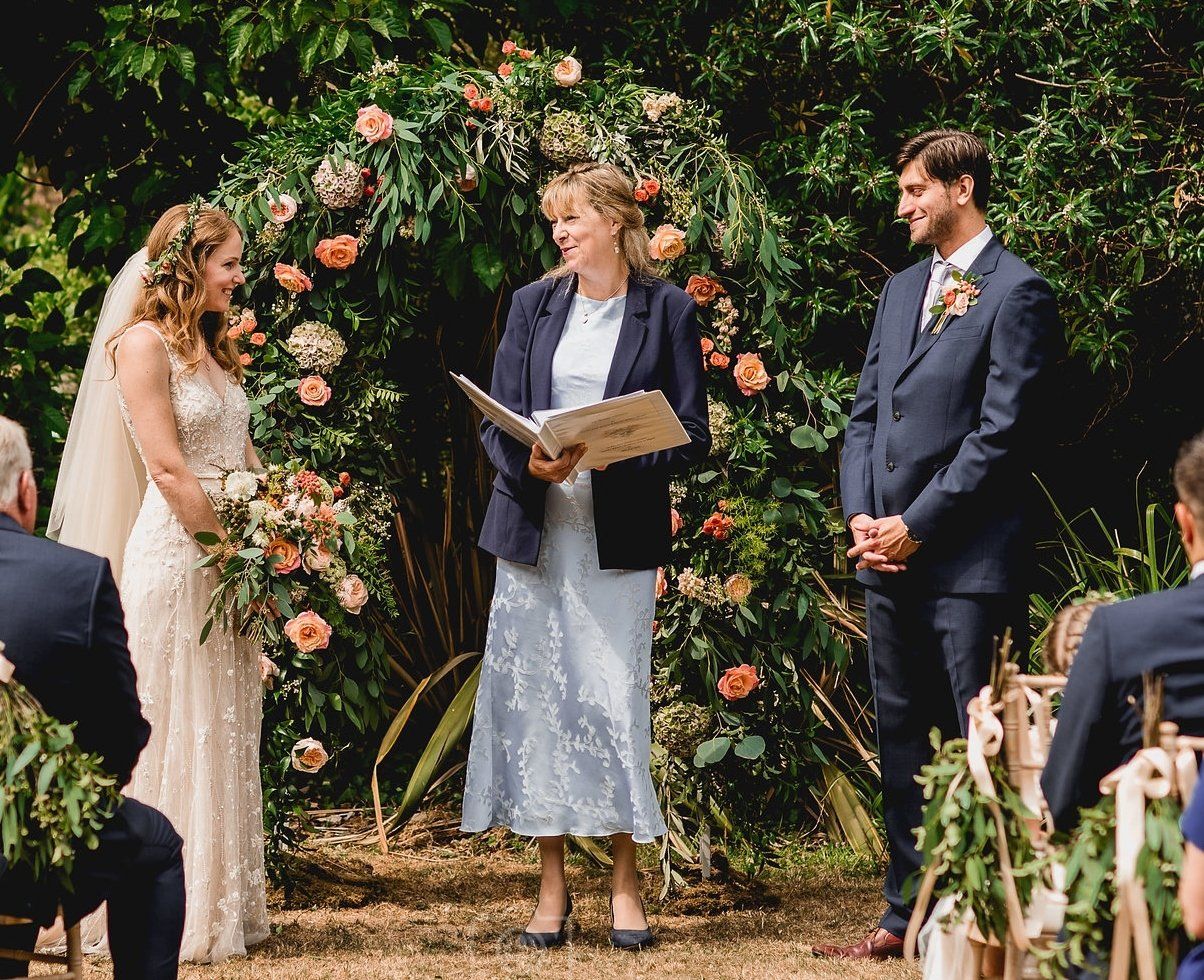 Wedding ceremony outdoors: Bride and groom facing officiant under floral arch; guests seated.