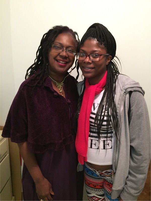 Woman with locs smiles next to a younger person with braids, both wearing glasses. They pose against a white wall.