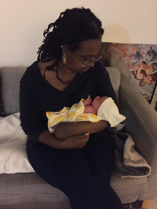 Woman holding a newborn baby wrapped in a yellow blanket, sitting on a gray couch, looking down.