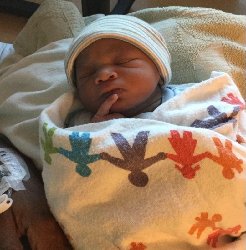 Newborn baby wrapped in a blanket, wearing a striped cap, and resting with a finger on its chin.