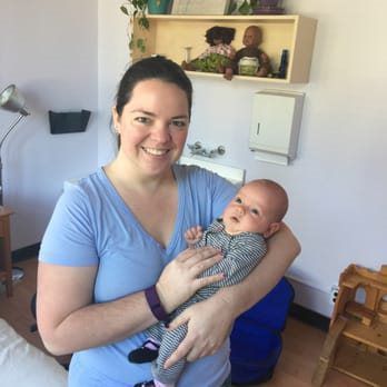 Woman in blue shirt holding a baby. Both smiling in a room with a sink and shelves.