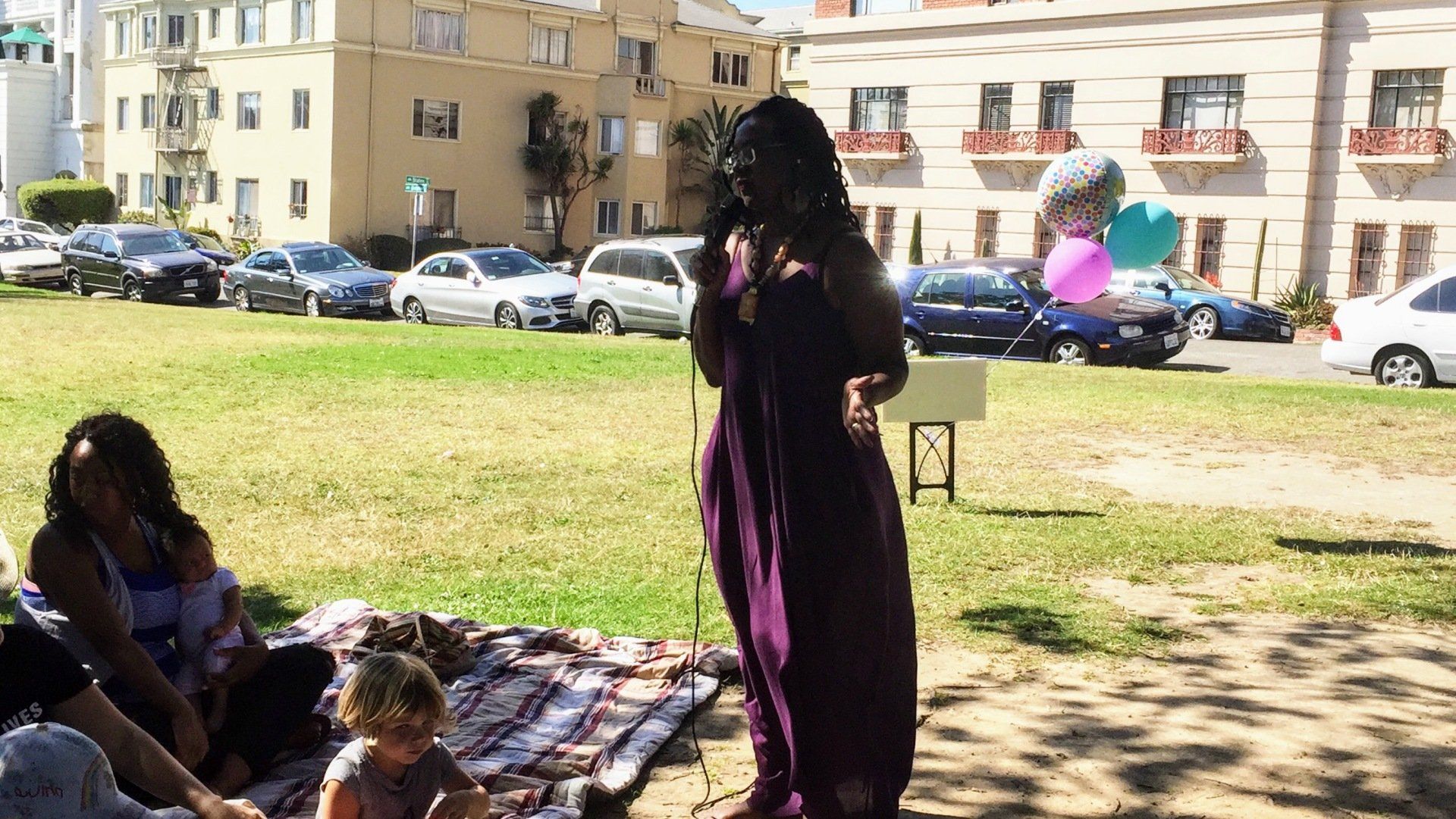 Woman speaking into a microphone in a park, balloons in the background, other people seated on a blanket.