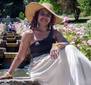 Woman in sun hat and one-shoulder top, sitting by a water feature in a garden, smiling.