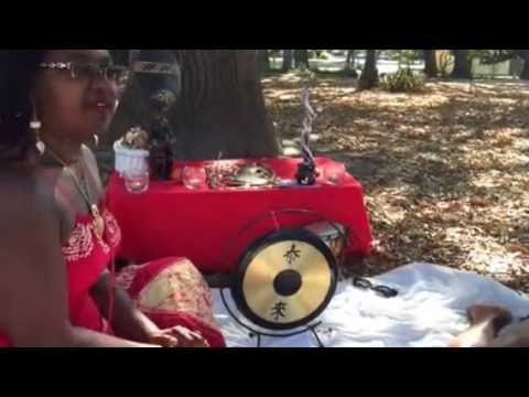 Woman in red dress near a table with gong, setting outdoors, possibly a spiritual gathering.