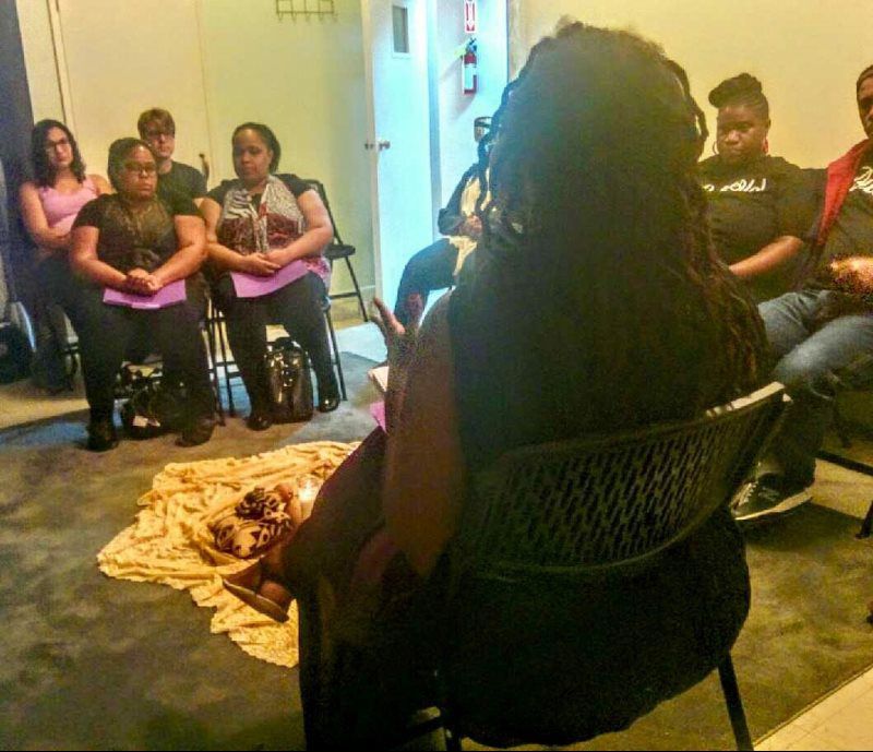 Group of people in a circle, some seated, with a lit candle and items on a cloth in the center, indoors.