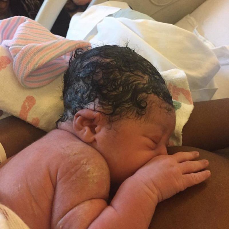 Newborn baby, dark hair, skin, resting on someone's chest, hospital setting.