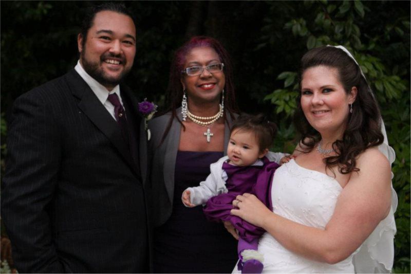 Wedding photo: Couple with a baby, and another person, all smiling in front of foliage.