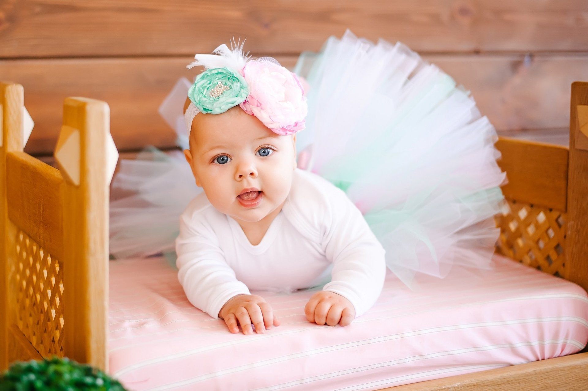 A baby girl wearing a tutu and headband is laying on her stomach in a wooden crib.