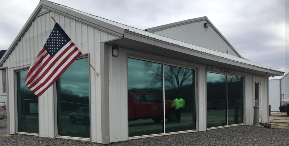 Small gray building with large windows and an American flag outside under a cloudy sky