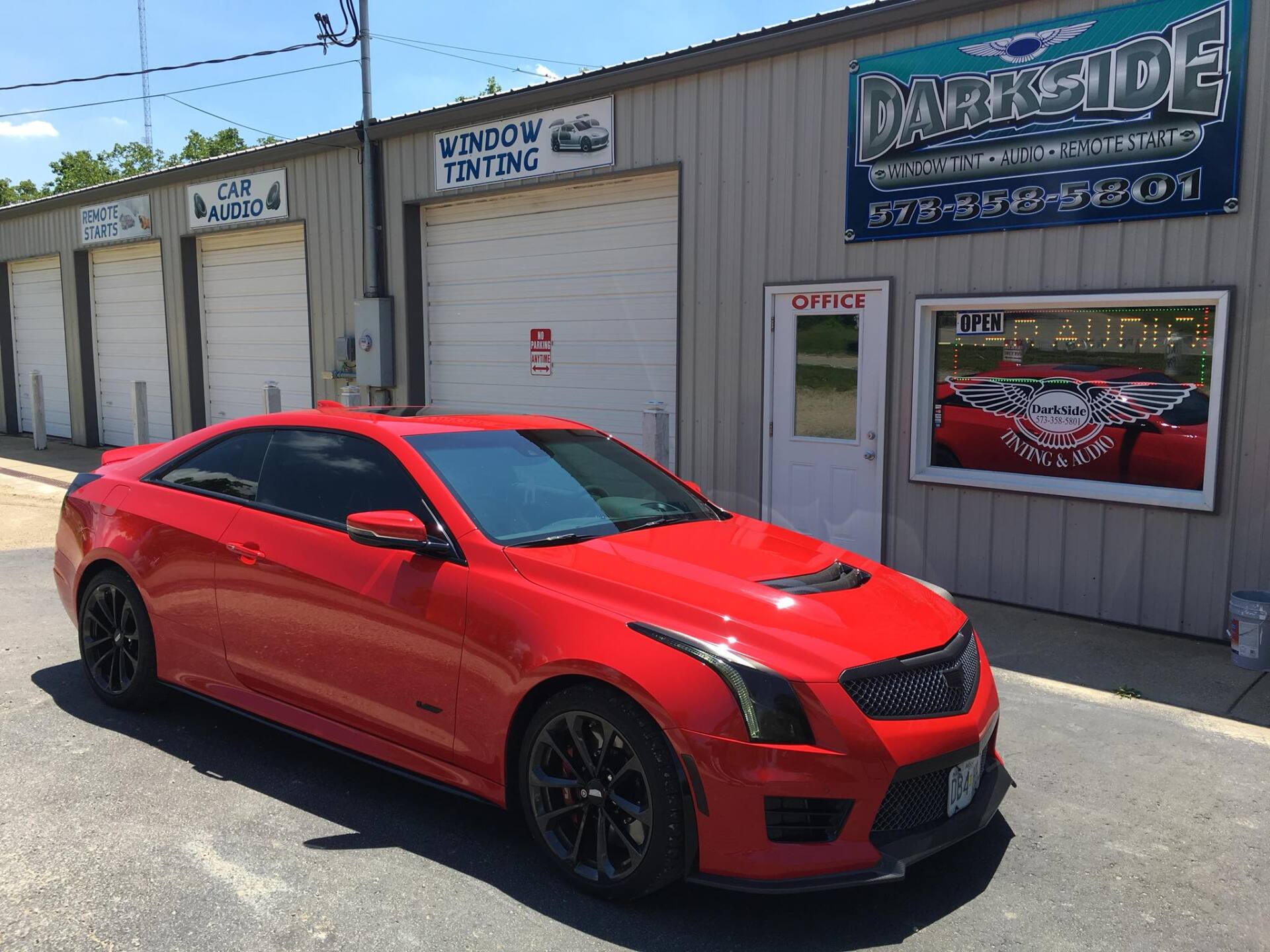 Red sports car parked outside an auto shop with garage doors and signage on a sunny day